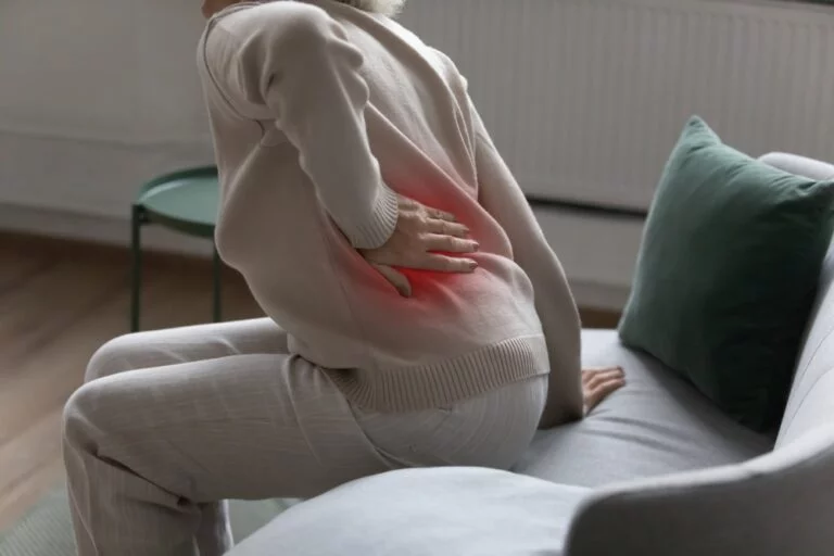 An older adult seated on a sofa holding their hip and lower back, with the painful area highlighted